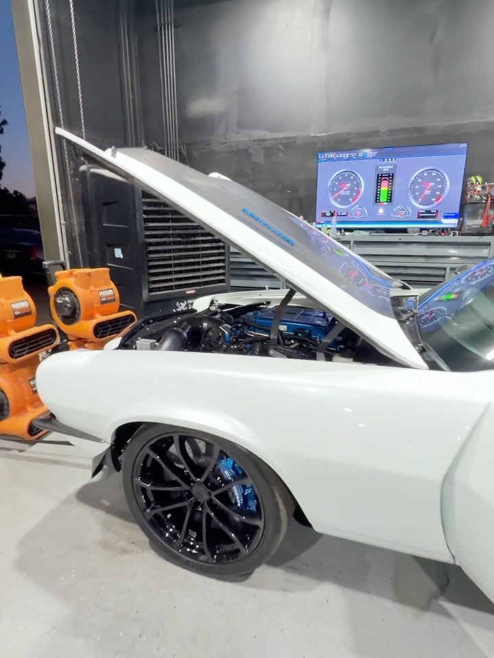 White Camaro with the hood up on the dyno at Mayne Designs, live gauges on the screen behind